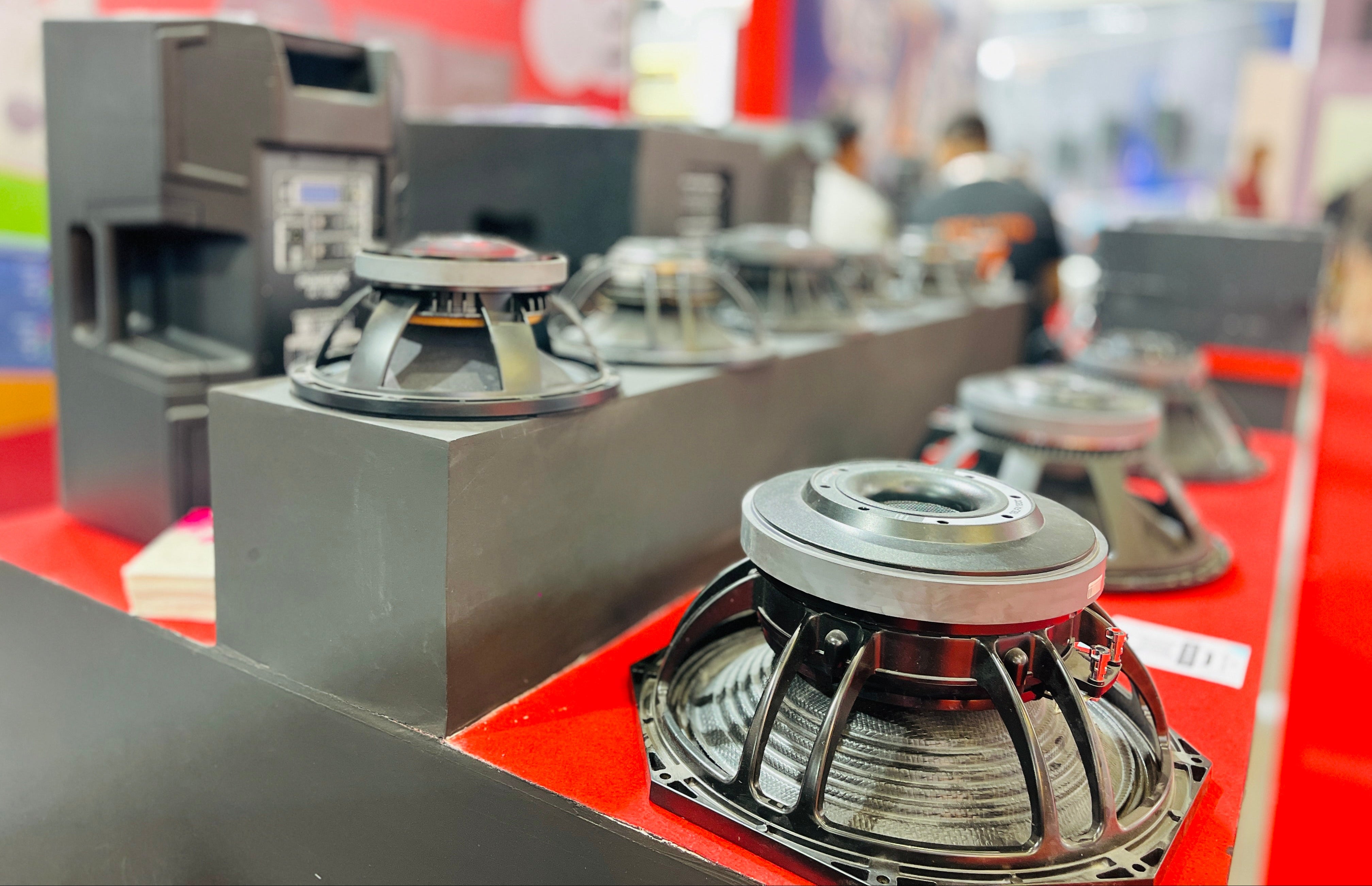 A row of large black and silver speaker drivers displayed on a red table at what appears to be a trade show or exhibition, with a blurred background showing booths and people.